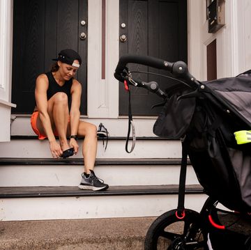 running postpartum woman sits on steps and puts her running shoes on while a stroller sits on the sidewalk